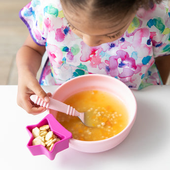 A child wearing a bright patterned bib enjoys soup from Bumkins Silicone Grip Bowl: Pink at a white table, using a spoon.