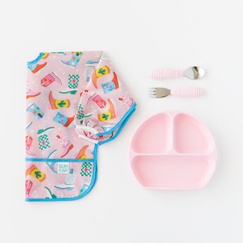Bumkins Spoon + Fork: pink rests near a pink Grip Dish and a colorful sleeved bib with a boot print, on a white background.