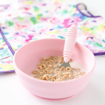 Bumkins Spoon + Fork: Pink and Bumkins Silicone Grip Bowl: Pink with cereal and milk, beside a colorful bib on a white background.