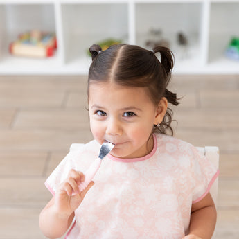 Child wearing a Bumkins Junior Bib: Floral, eating from a Bumkins Spoon + Fork: Pink.