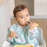 A toddler in a rainbow bib sits in a high chair using Bumkins Spoon + Fork: Pineapple, looking up at the camera.
