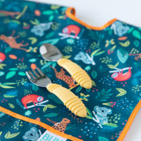 Bumkins Spoon + Fork: Pineapple set for self-feeding sits on a vibrant animal-print bib with koalas, sloths, and leaves.