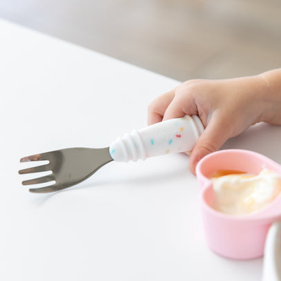 A childs hand grips a Bumkins Spoon + Fork: Vanilla Sprinkle, featuring a white silicone handle with colorful speckles. Beside it sits a pink cup of yogurt on a white table, ideal for encouraging self-feeding.