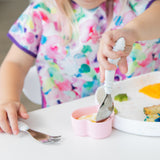 A child in a floral bib enjoys colorful food from a plate with Bumkins Spoon + Fork: Vanilla Sprinkle.