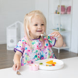 A smiling blonde girl in a floral bib uses Bumkins Spoon + Fork: Vanilla Sprinkle to eat from a white plate at a white table.