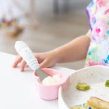 Close-up of a child using Bumkins Spoon + Fork: Vanilla Sprinkle to enjoy yogurt from a pink heart-shaped cup at a white table.