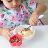 A child happily eats ice cream and sprinkles from a bowl with Bumkins Spoon + Fork: Vanilla Sprinkle at a white table.