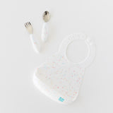 Bumkins Spoon + Fork: Vanilla Sprinkle sit near a matching bib on a white background.