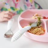 Close-up of a child's hand near a pink plate with pasta and veggies, and Bumkins Spoon + Fork: Vanilla Sprinkle on a white table.
