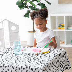 A girl paints pink at a table covered with a Bumkins Splat Mat: Hearts. A jar and art supplies are on the easy-wipe surface.