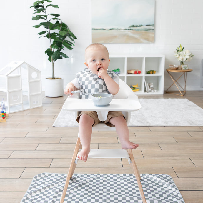 A baby sits in a high chair with a gray and white checkered bib, eating from a bowl. The room has wooden floors, a small white table, toys, and a painting on the wall. A potted plant and decorative items are in the background.