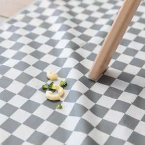 Tiny pasta and broccoli sit on a Bumkins Splat Mat: Charcoal Check, under a wooden table leg on the floor.