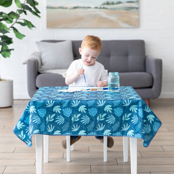 A child paints on a table covered by a Bumkins Splat Mat: Blue Tropic in front of a gray sofa and plant in a bright living room.