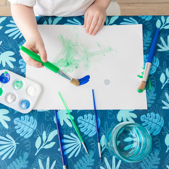 A child paints with green and blue paints on white paper on a Bumkins Splat Mat: Blue Tropic.