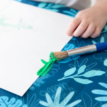 Close-up of a child's hand and a paintbrush with green paint near a white paper on a Bumkins Splat Mat: Blue Tropic.