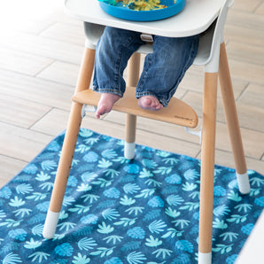 A baby in jeans sits barefoot in a high chair atop a Bumkins Splat Mat: Blue Tropic on a wood floor.