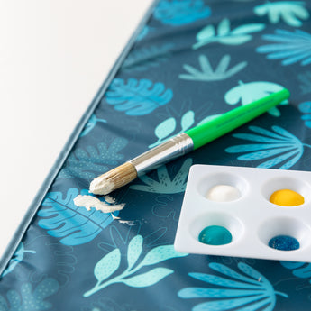 Close-up of Bumkins Splat Mat: Blue Tropic on a white background with a paint palette and green paintbrush on top.
