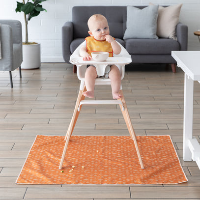 A baby sits in a high chair wearing a yellow bib, with a bowl in front. The high chair is on a wooden floor, and an orange mat with a pattern is beneath it. A gray sofa and cushions are in the background, along with a potted plant.