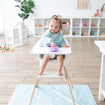 A toddler in a high chair eats from a purple bow in a playroom, with a Bumkins Splat Mat: Rainbows protecting the floor below.