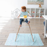 A toddler in a high chair smiles, wearing a bib, with a bowl and spoon on the Bumkins Splat Mat: Rainbows in a bright playroom.