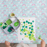 A child paints green dots on paper with a palette, using the Bumkins Splat Mat: Rainbows as a pastel tablecloth.