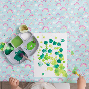 On a Bumkins Splat Mat: Rainbows, a child paints green dots on paper with a silicone plate of paints and art supplies nearby.