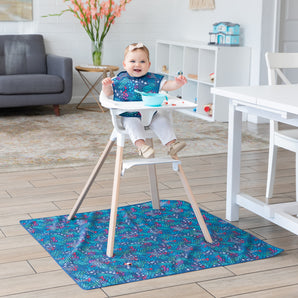 A baby in a highchair smiles wearing a blue bib with a bowl on tray, and a Bumkins Splat Mat: Animal Prints underneath in the bright room.