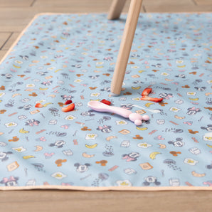 Bumkins Splat Mat: Mickey + Minnie Sunny Side Up lays on a wood floor under a wooden chair leg, with strawberry pieces and a spoon on top.