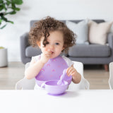 A toddler with curly hair sits at a table, wearing a Bumkins Silicone Bib 2 Pack: Vanilla Sprinkle and Rainbows and eating from a purple bowl.