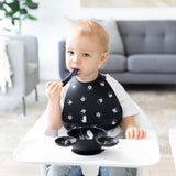 A toddler in a high chair enjoys baby-led weaning with Bumkins Silicone Feeding 3-Piece Set: Mickey Mouse Faces B+W in a bright living room.