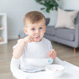 A young child wearing a Bumkins Silicone Bib 2 Pack: Vanilla Sprinkle and Hearts eats from a matching bowl in a high chair.