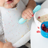 Close-up of a baby in a Bumkins Silicone Bib: Vanilla Sprinkle eating messily with a blue spoon, surrounded by food and a bowl on the table.