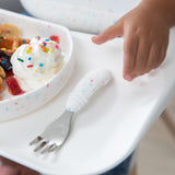Close-up of a child's hand reaching for a Bumkins Spoon + Fork: Vanilla Sprinkle next to a matching plate with waffles on a high chair tray.