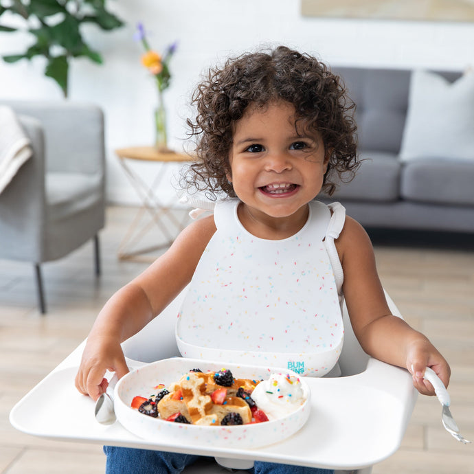 A smiling girl with curly hair sits in a high chair, wearing a Bumkins Silicone Bib: Vanilla Sprinkle next to a plate of waffles and berries.