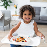 A smiling girl with curly hair sits in a high chair, wearing a Bumkins Silicone Bib: Vanilla Sprinkle next to a plate of waffles and berries.