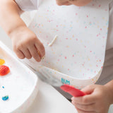 Close-up of a child wearing a Bumkins Silicone Bib: Vanilla Sprinkle, placing food into the pocket while holding a red fork.