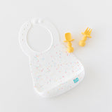Bumkins Silicone Bib: Vanilla Sprinkle rests on a white background next to a pair of yellow baby utensils.