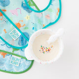 A bowl of white baby food with sprinkles sits beside Bumkins Starter Bib 2 Pack: Ocean Life & Whale Tail, waterproof bibs.