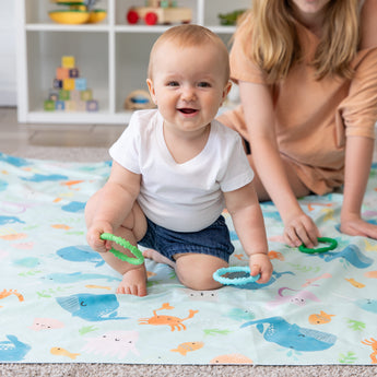 A baby on a sea creature blanket holds Bumkins Silicone Teething Rings 4 Pack: Summer, while a woman in a peach shirt sits nearby.