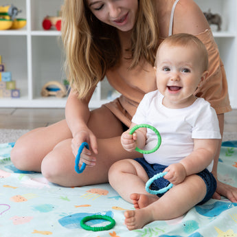A baby plays with Bumkins Silicone Teething Rings 4 Pack: Summer on a blanket as a woman holds another, with toys on shelves in the background.
