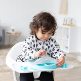 A young girl wearing a black & white bib in a high chair lifts the lid of Bumkins Grip Dish Essential Infant 4-Piece Mealtime Set: Blue.