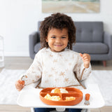 In a high chair, a curly-haired child wearing a Bumkins Sleeved Bib: Palm Daze smiles while holding a spoon next to a divided plate of food.
