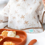 Close-up of a child in a Bumkins Sleeved Bib: Palm Daze enjoying baby-led weaning with an orange plate of crackers and dip in the foreground.