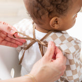 An adult ties the brown ribbon closure on a Bumkins Sleeved Bib: Sand Check as a baby in a white shirt sits ready for baby-led weaning.