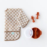 Bumkins Sleeved Bib: Sand Check lays folded next to an orange bowl & spoon set and orange toddler utensils on a white background.