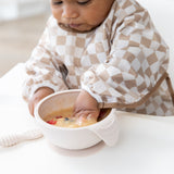 A baby wearing Bumkins Sleeved Bib: Sand Check eagerly explores porridge, embracing baby-led weaning at a white table.