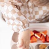 Close-up of a child in a Bumkins Sleeved Bib: Sand Check reaching for strawberries and oranges, embracing baby-led weaning.