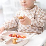 A toddler in a Bumkins Sleeved Bib: Sand Check enjoys baby-led weaning with fruit from a tan divided plate in a high chair.