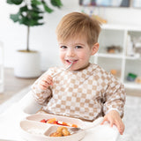 A happy toddler in a Bumkins Sleeved Bib: Sand Check sits in a high chair, enjoying baby-led weaning with a spoon from a beige plate.