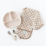 Bumkins Sleeved Bib: Sand Check is paired with a divided plate and matching spoon and fork on a white background.